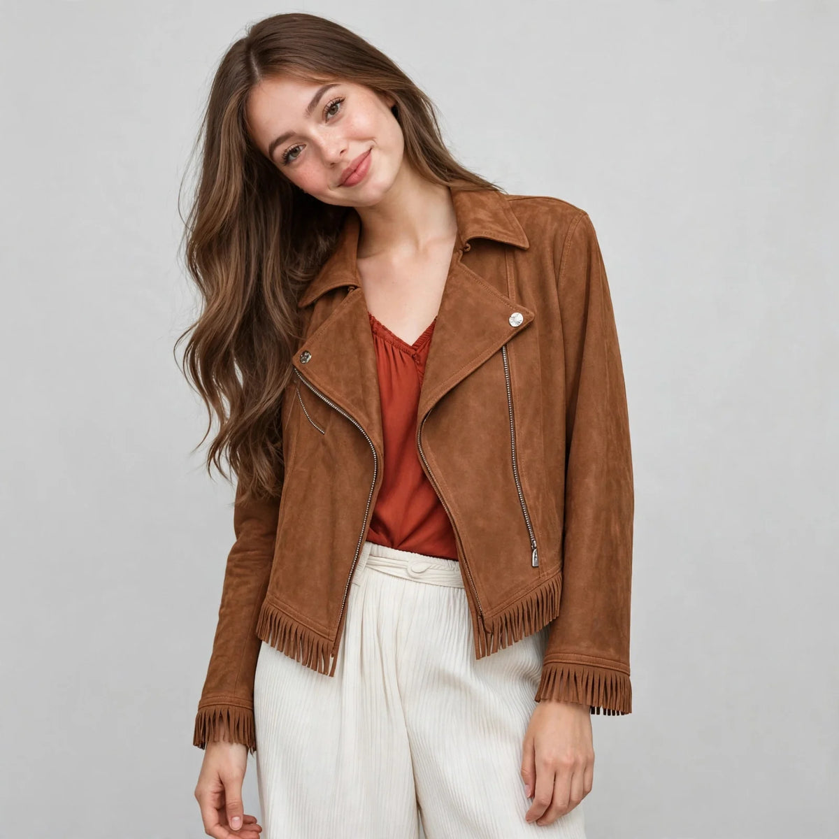 Woman standing in a caramel brown faux suede fringe moto jacket over a rust top and white pants, gray studio backdrop.