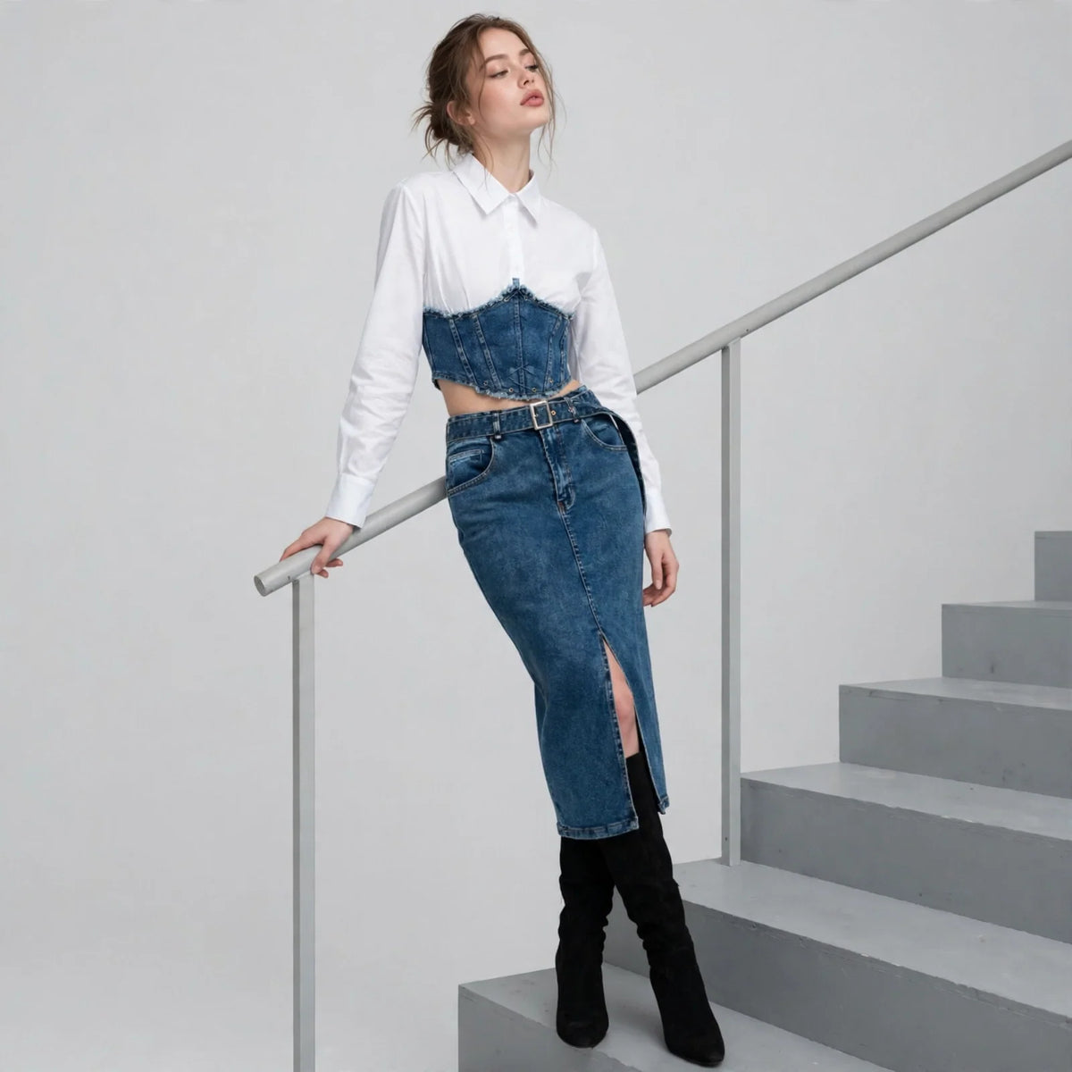 Woman leaning on stair railing in white shirt, denim corset top, dark denim slit midi skirt, black boots, light backdrop.