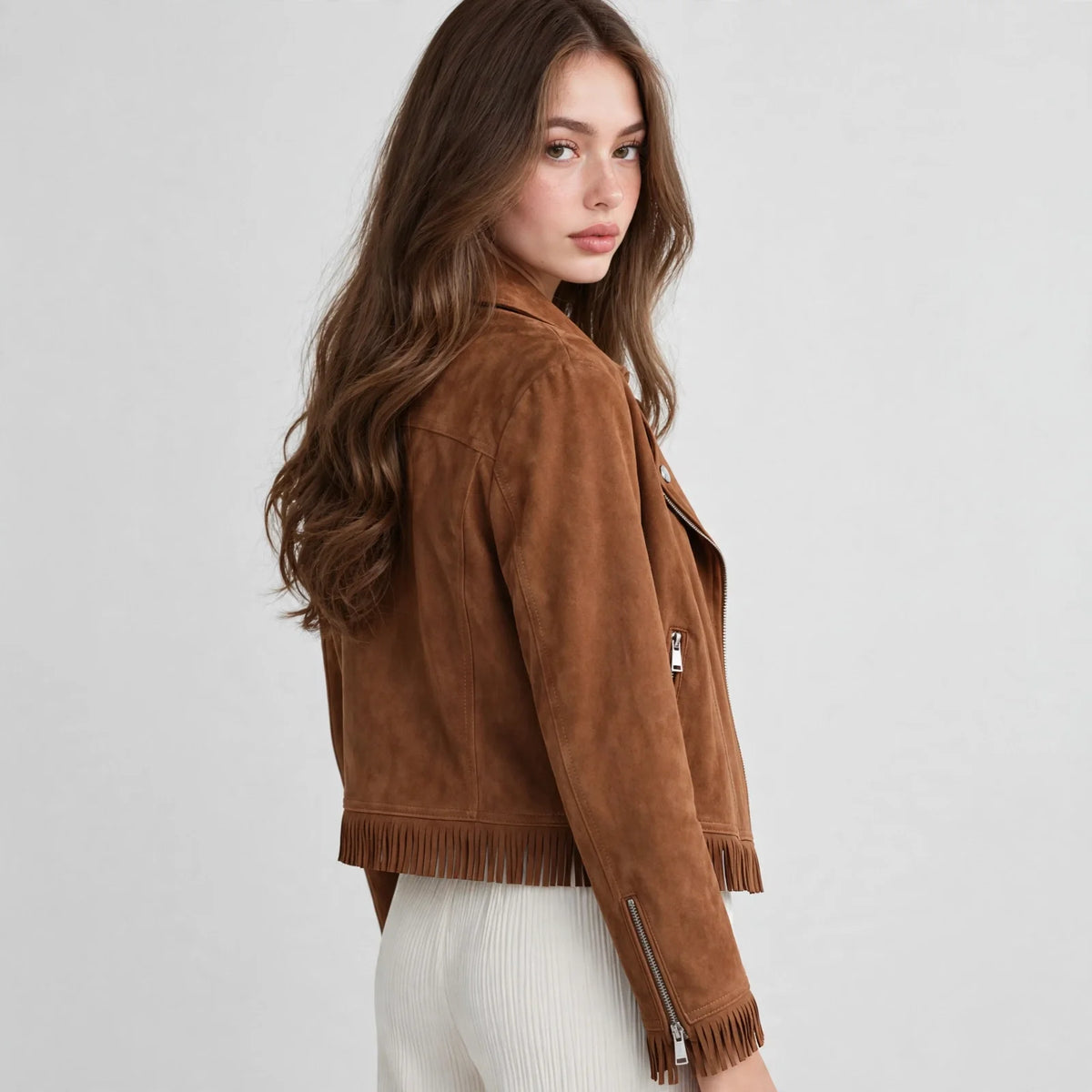 Three-quarter back view of a woman in a caramel brown faux suede fringe moto jacket and white pants, gray studio backdrop.