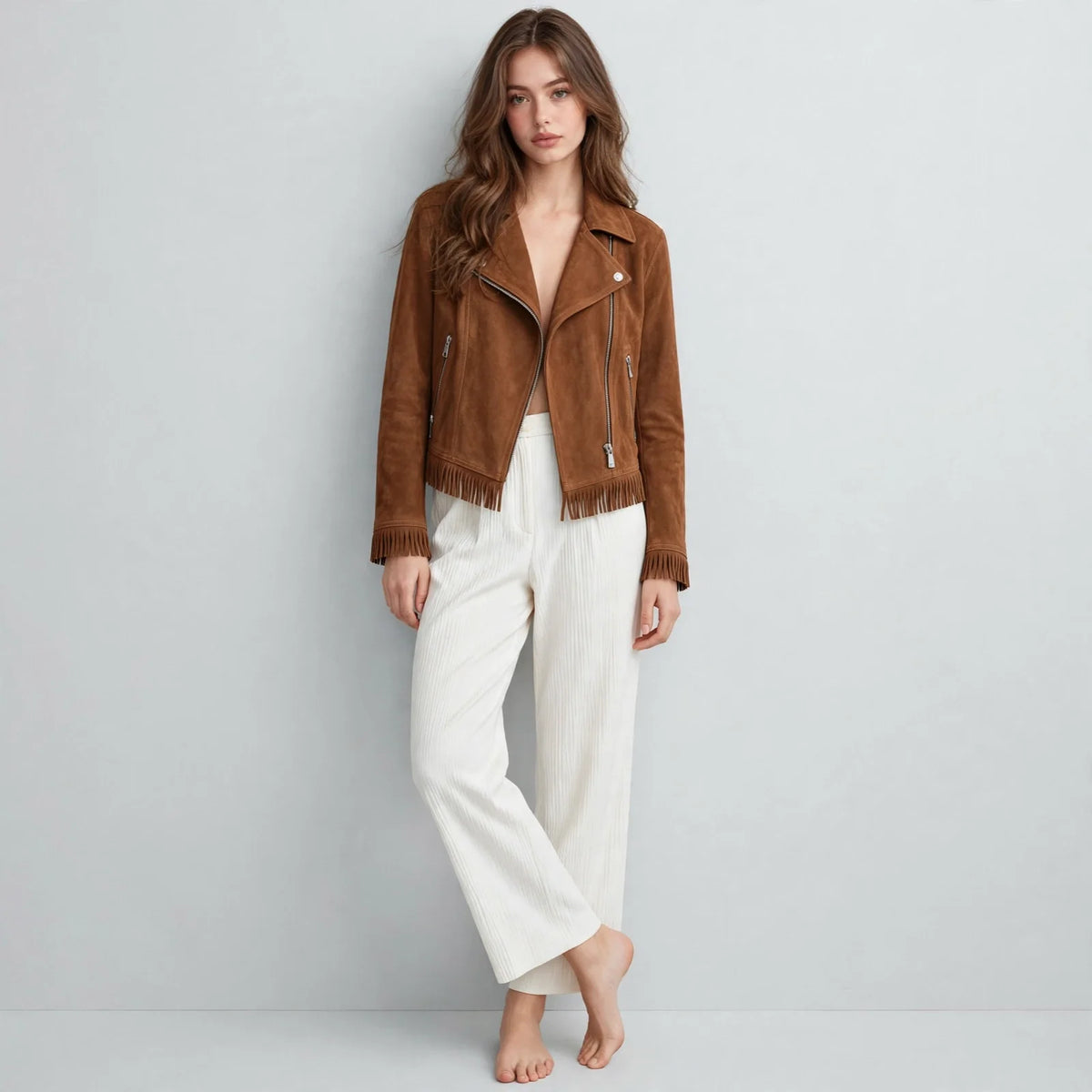 Full-length front view of a woman in a caramel brown faux suede fringe moto jacket and white pants, barefoot, gray studio.