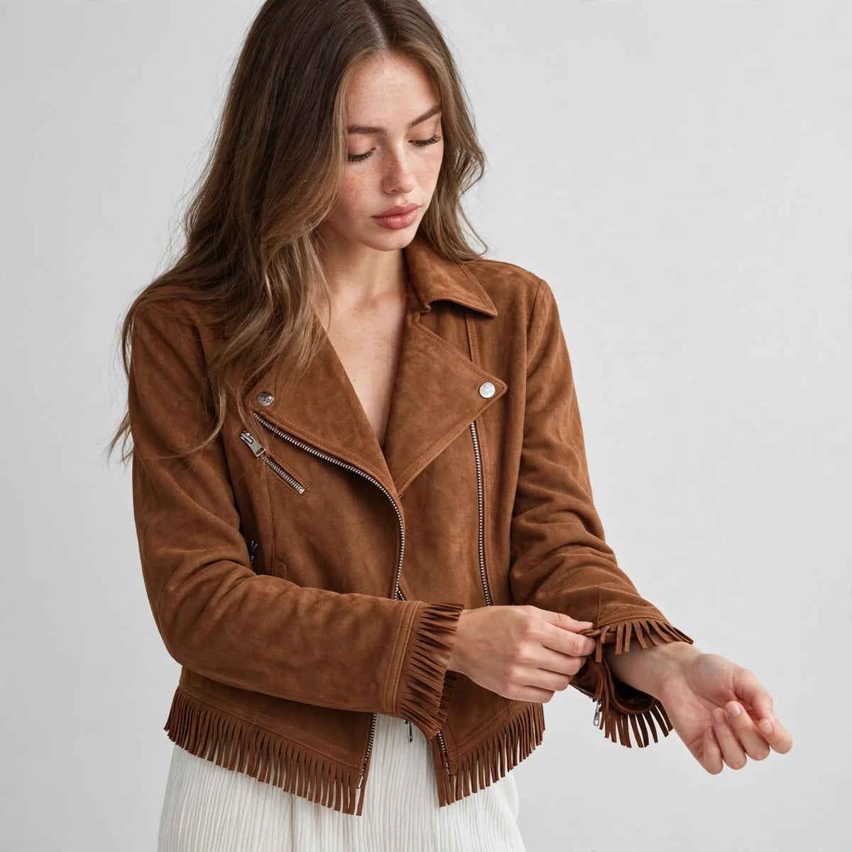 Woman adjusting her sleeve in a caramel brown faux suede fringe moto jacket over a white top and jeans, gray studio backdrop.