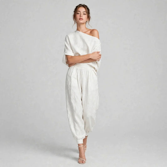 Woman standing with arms crossed in a white one-shoulder top and matching loose joggers, light gray studio background.