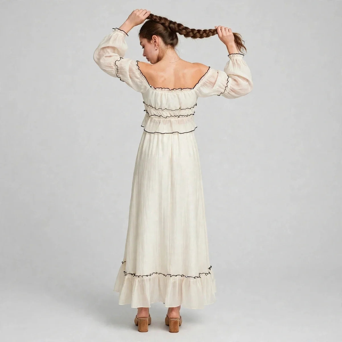 Back view of a woman holding her braid in a cream off-shoulder top and long ruffled skirt set with black trim, gray studio.