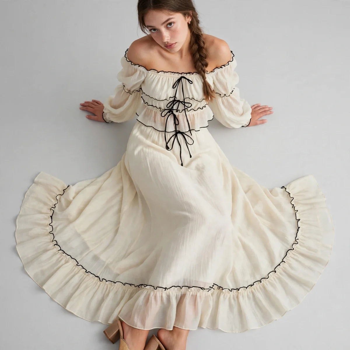 Woman seated on the floor with skirt spread in a cream off-shoulder top and ruffled skirt set with black trim, gray studio.