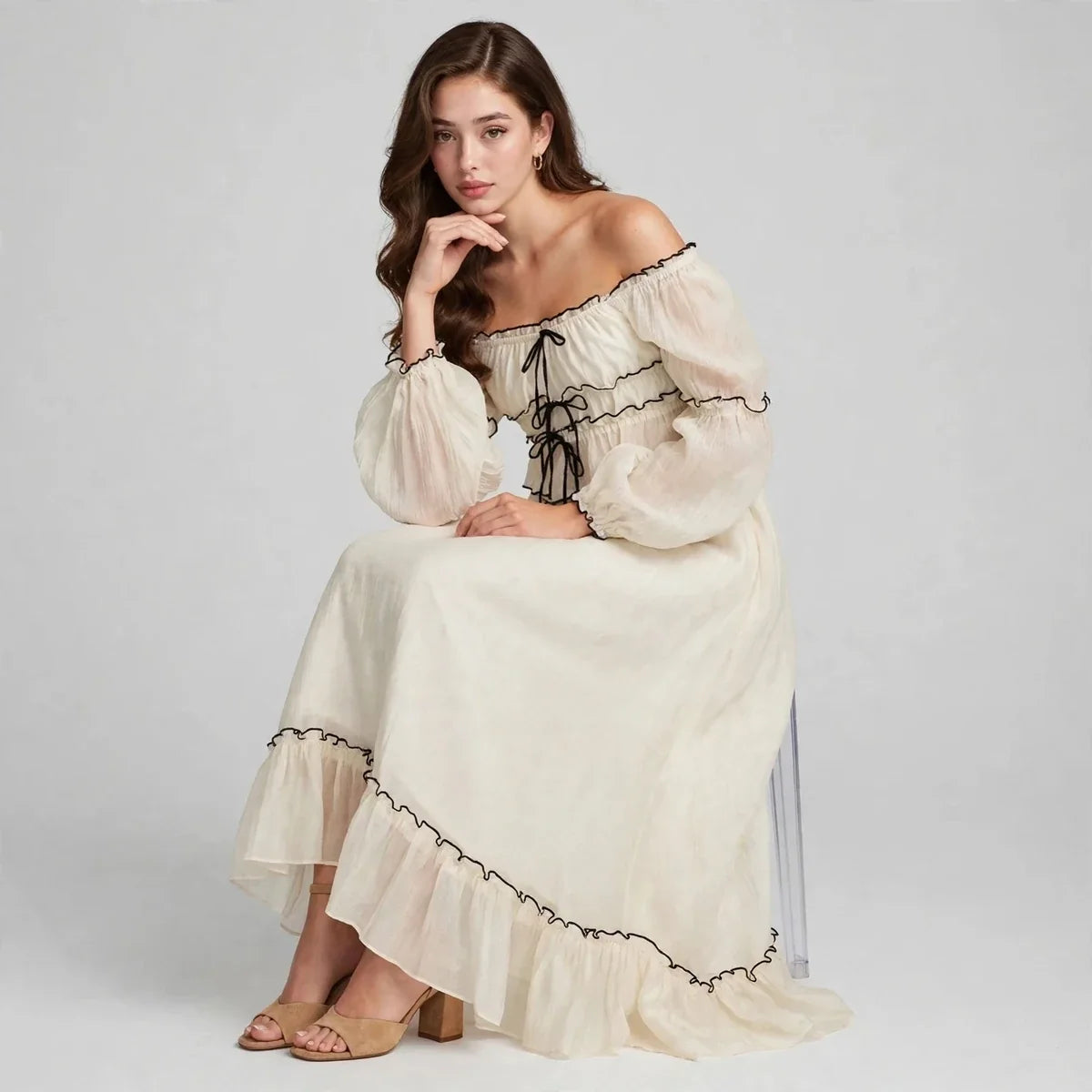 Woman seated on a chair with hand near chin in a cream off-shoulder top and ruffled long skirt set, gray studio backdrop.