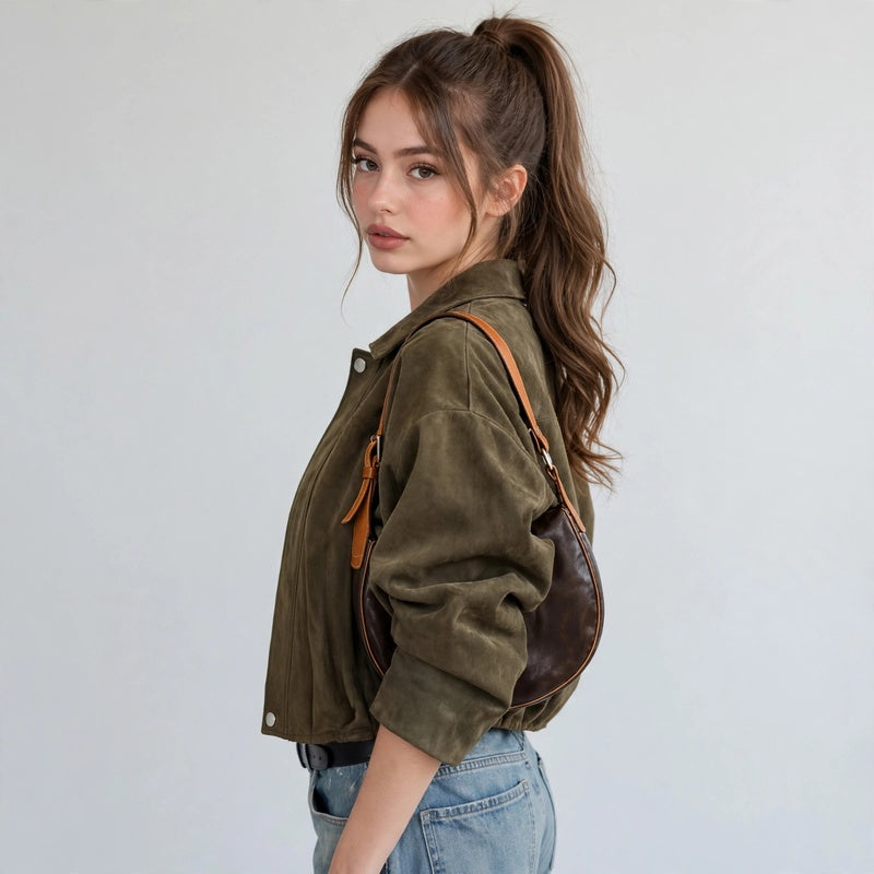 Side view of a woman wearing an olive green cropped suede bomber jacket with blue jeans and a brown shoulder bag.