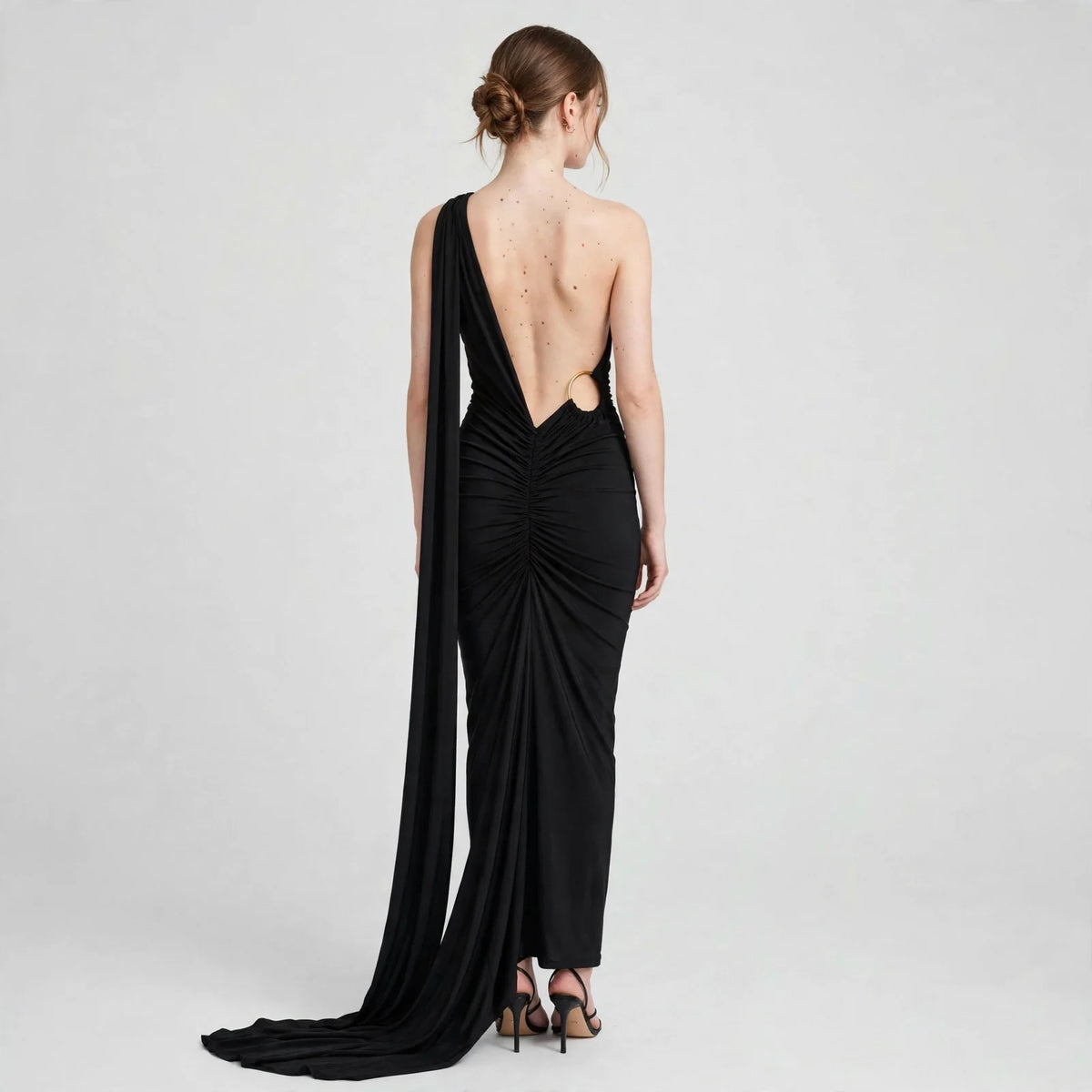 Woman modeling a black one-shoulder evening gown from the back with a deep open back and long train, gray background.