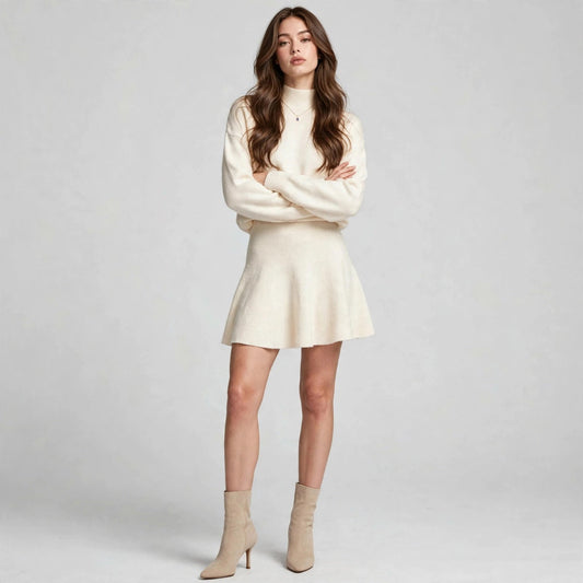 Woman in a cream turtleneck knit sweater and matching flared mini skirt set with beige ankle boots, gray studio background.