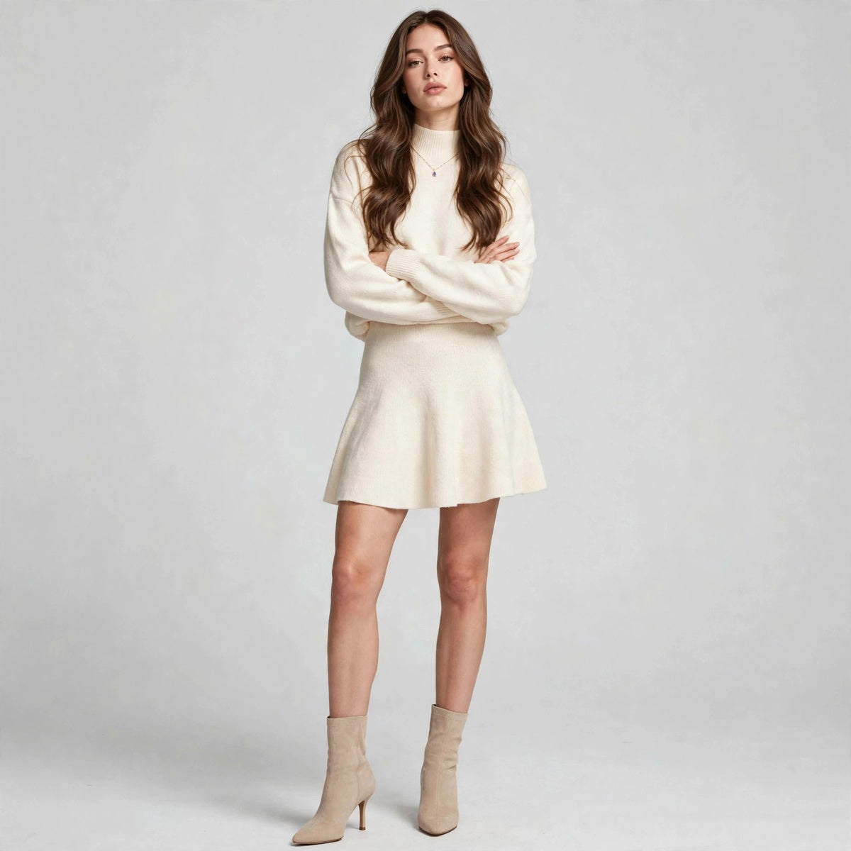 Woman in a cream turtleneck knit sweater and matching flared mini skirt set with beige ankle boots, gray studio background.
