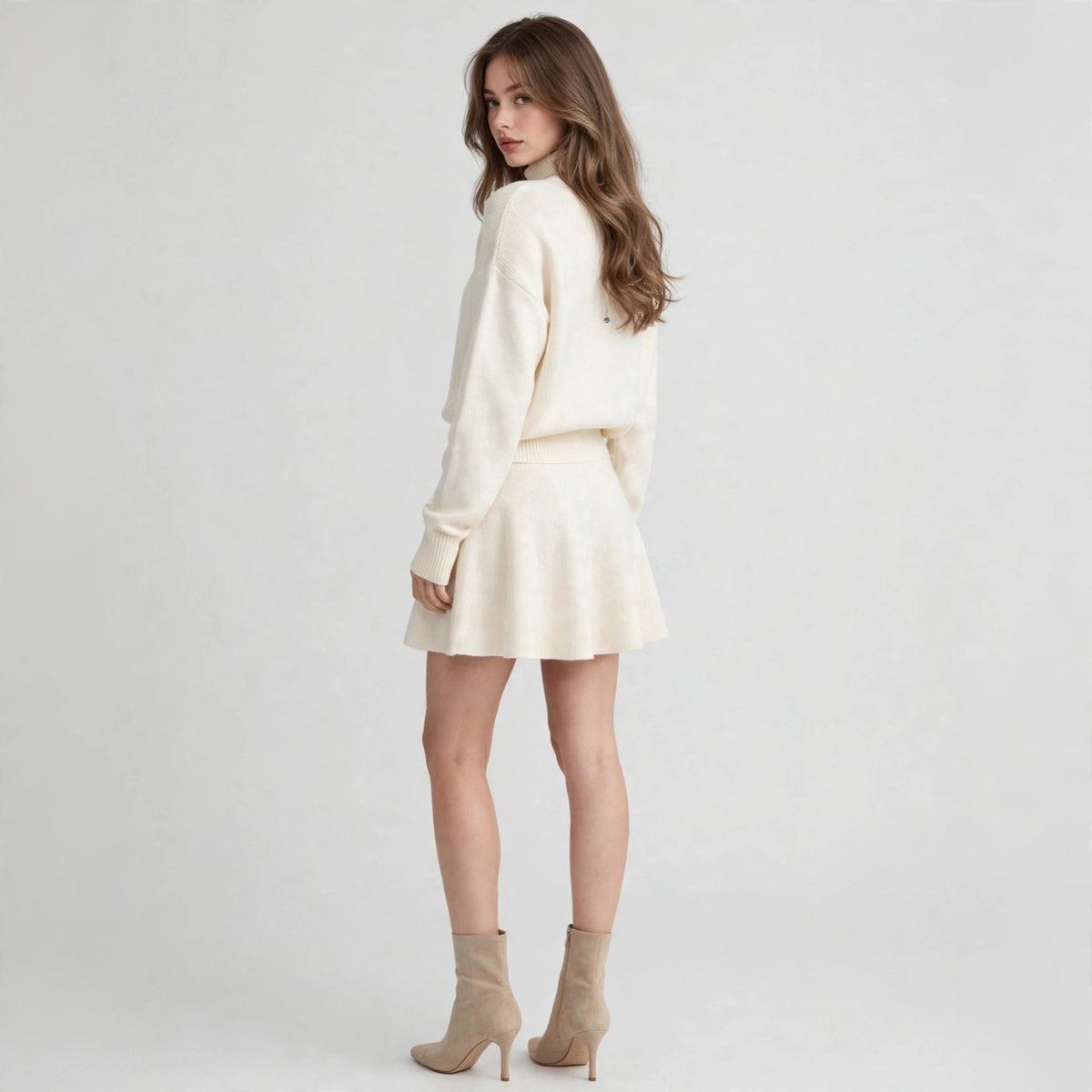 Back three-quarter view of a cream turtleneck sweater and flared mini skirt set with beige ankle boots, gray studio.