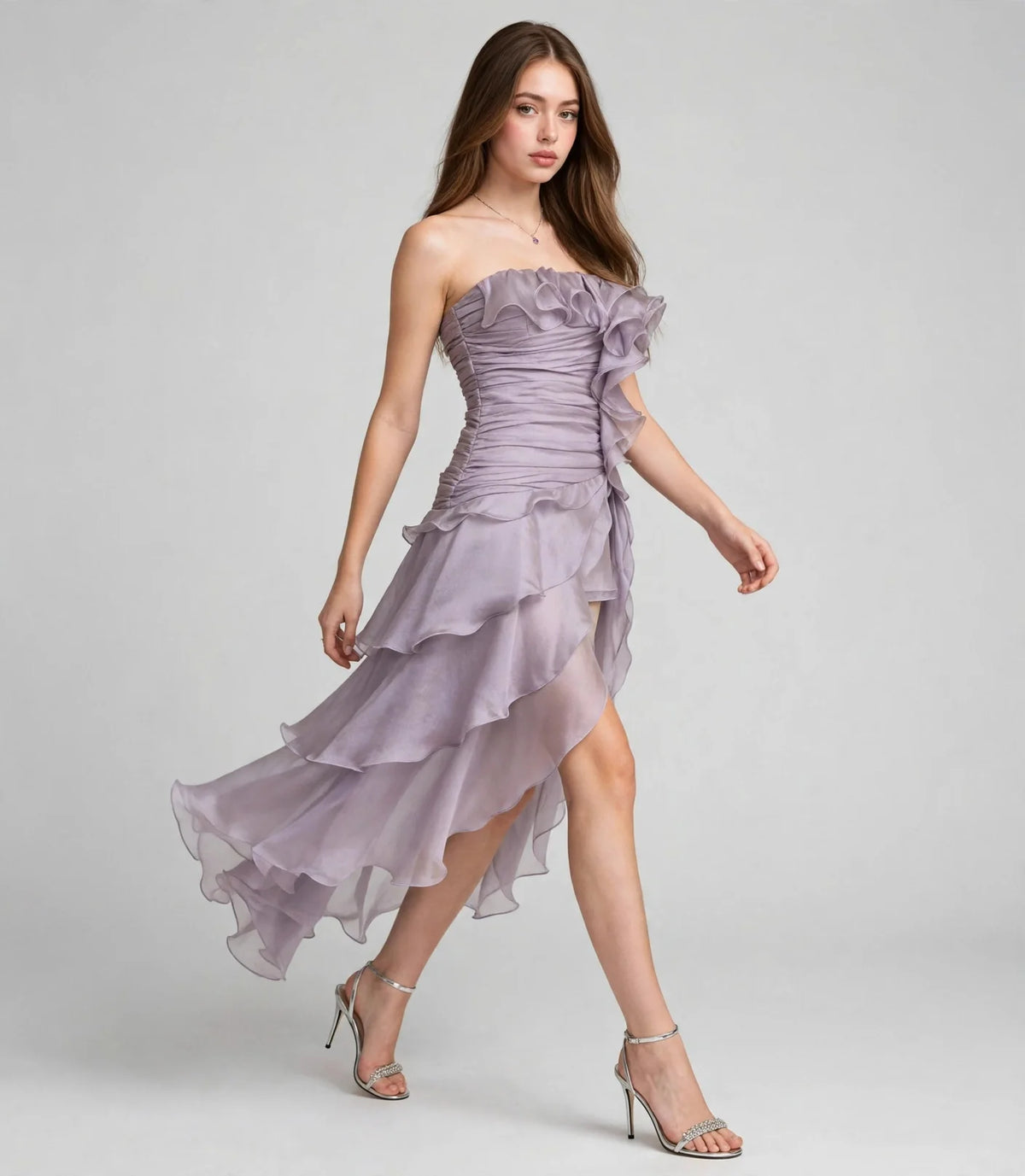 Woman walking in a lavender chiffon strapless dress with ruffled tiered skirt and silver heels, gray studio background.