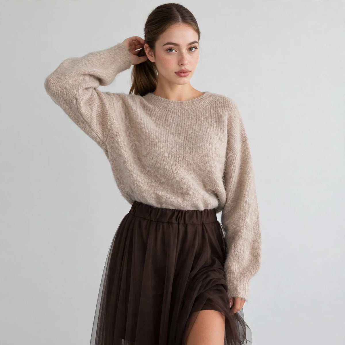 Model in beige fuzzy dropped-shoulder sweater with hand in hair, paired with dark tulle skirt, front view, studio shot.