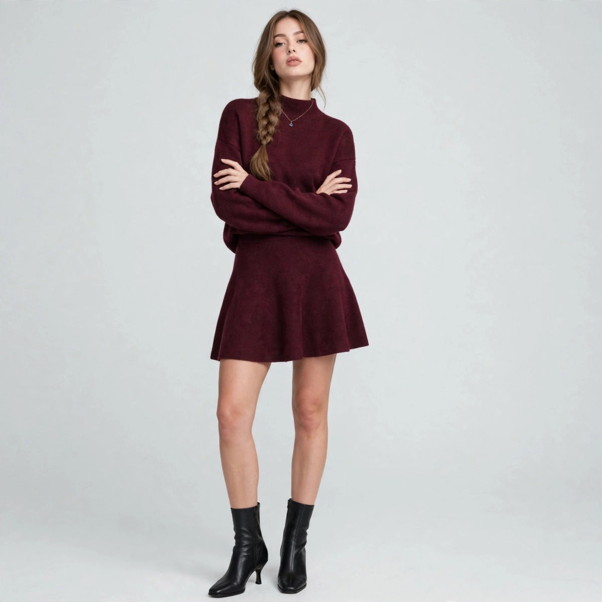 Woman in a burgundy mock-neck knit sweater and flared skirt set, arms crossed, black ankle boots, gray studio background.
