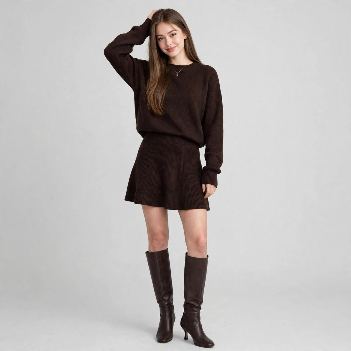 Woman in a dark brown knit sweater and mini skirt set gesturing, brown knee-high boots, gray studio background.