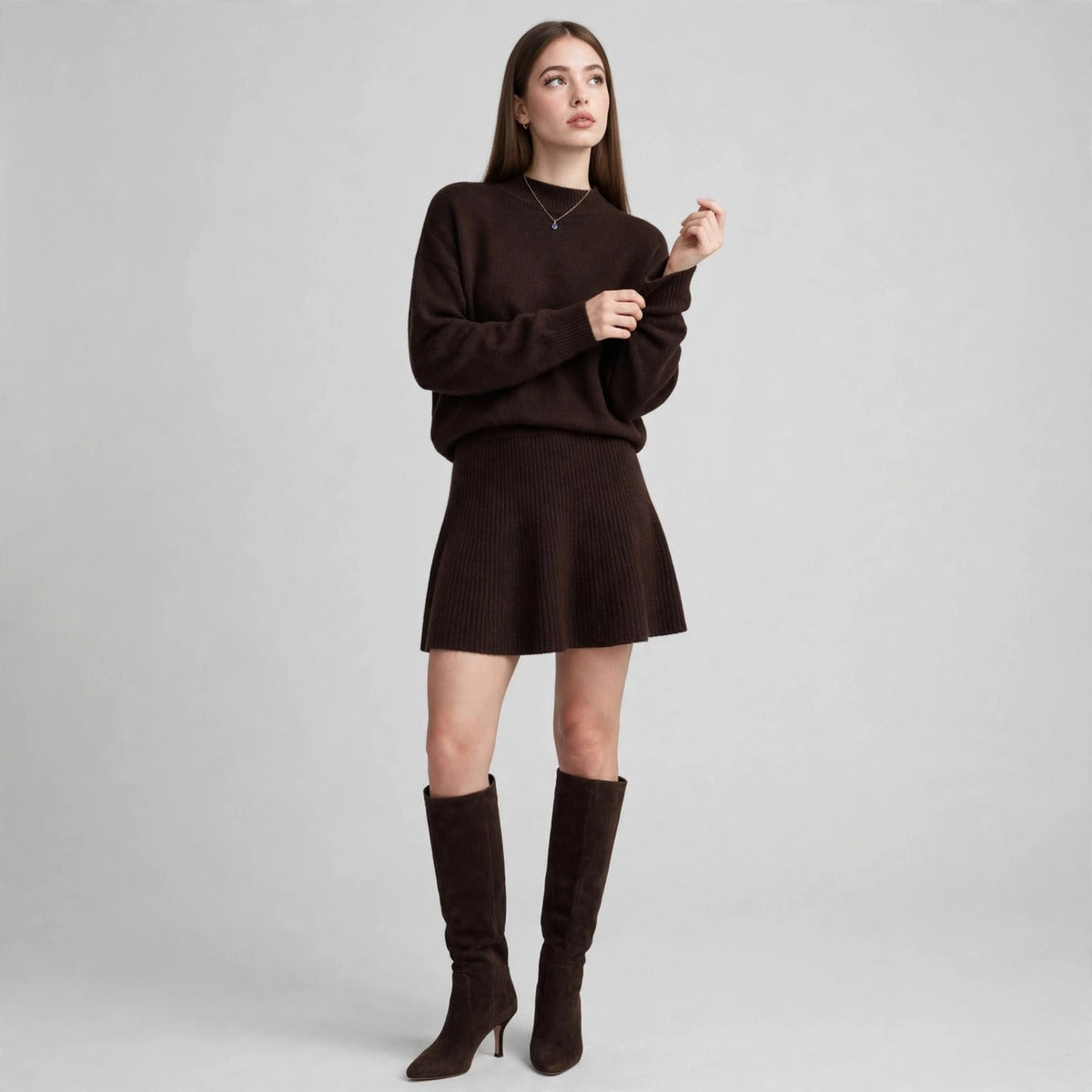 Woman in a dark brown ribbed knit sweater and matching mini skirt set with brown knee-high boots, gray studio.