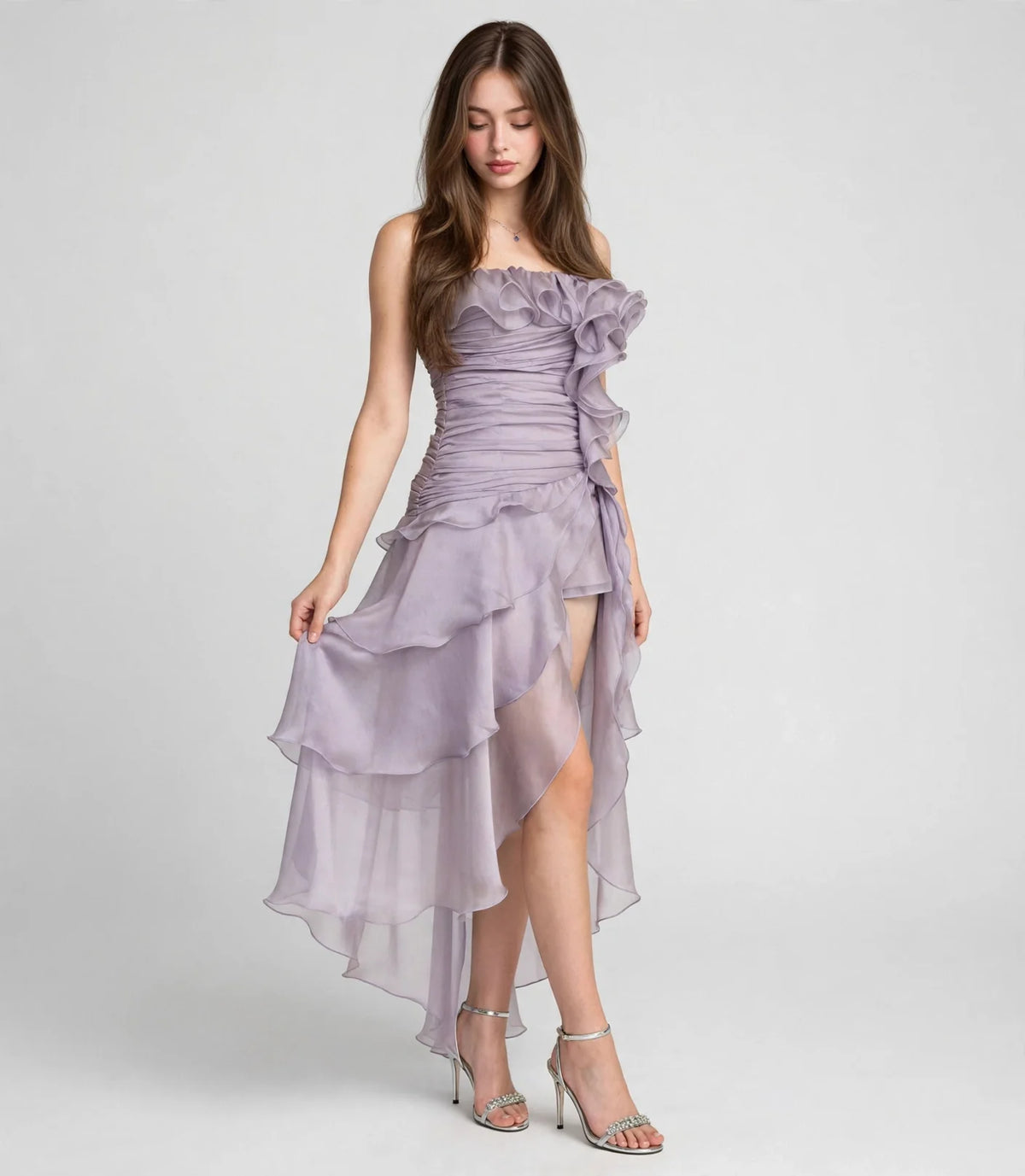 Woman standing in a lavender strapless ruffled dress with a high-low tiered skirt, front view, light gray studio background.