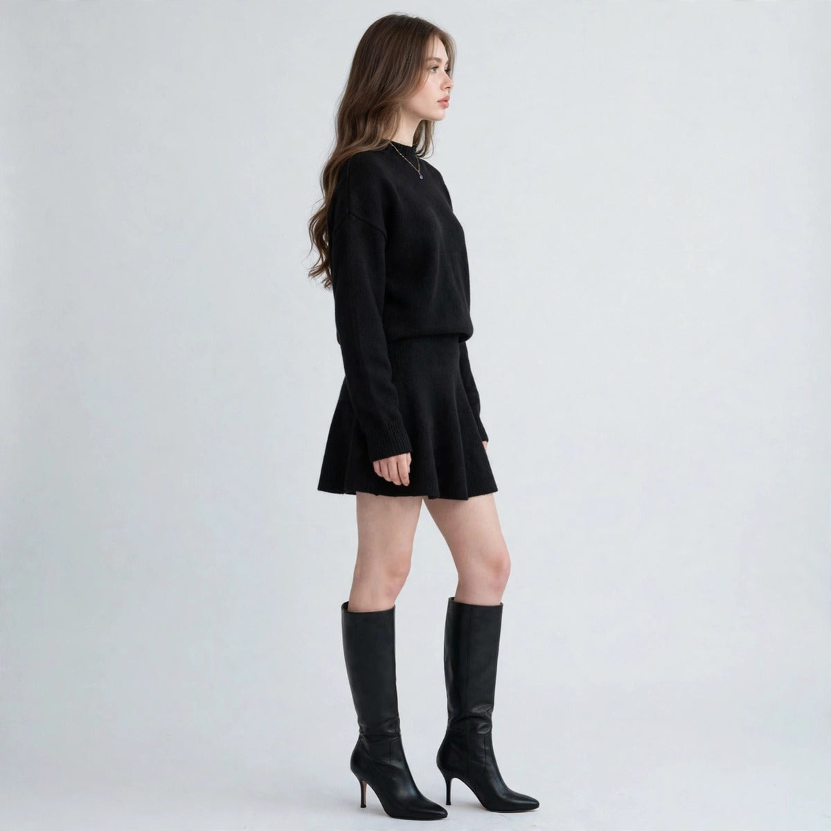 Side profile of a black knit sweater and matching mini skirt set with black knee-high boots, gray studio background.