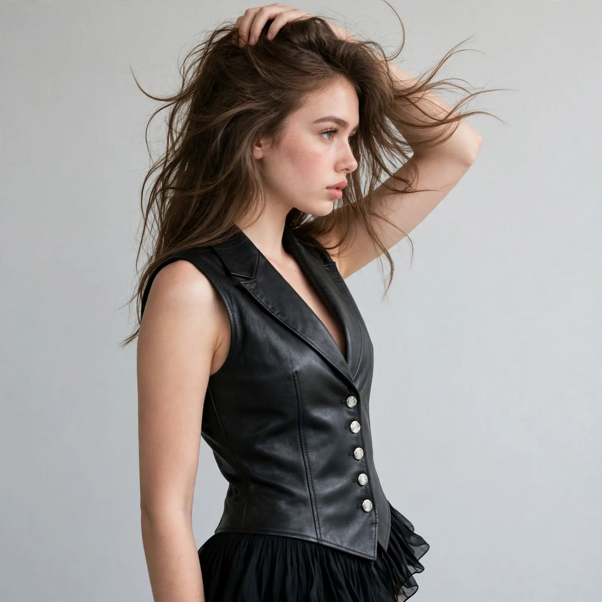 Model with hand in hair wearing a black faux leather button-down vest coat with lapel collar, wind in hair, studio.