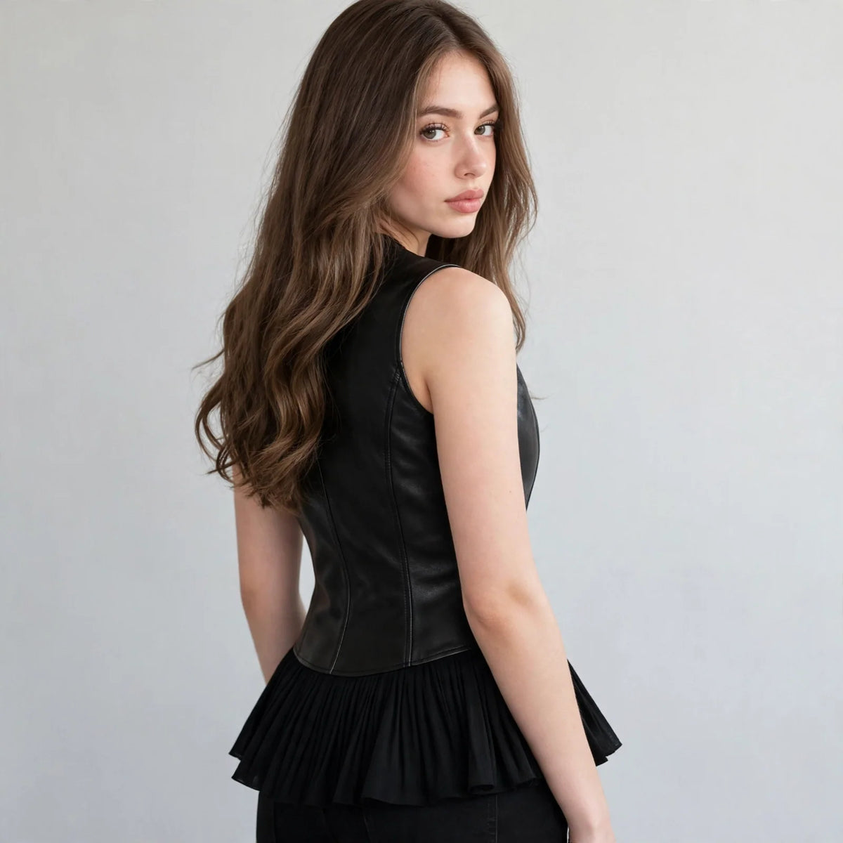 Back view of a black sleeveless vest coat with peplum ruffle hem on a model with long wavy hair, gray studio backdrop.