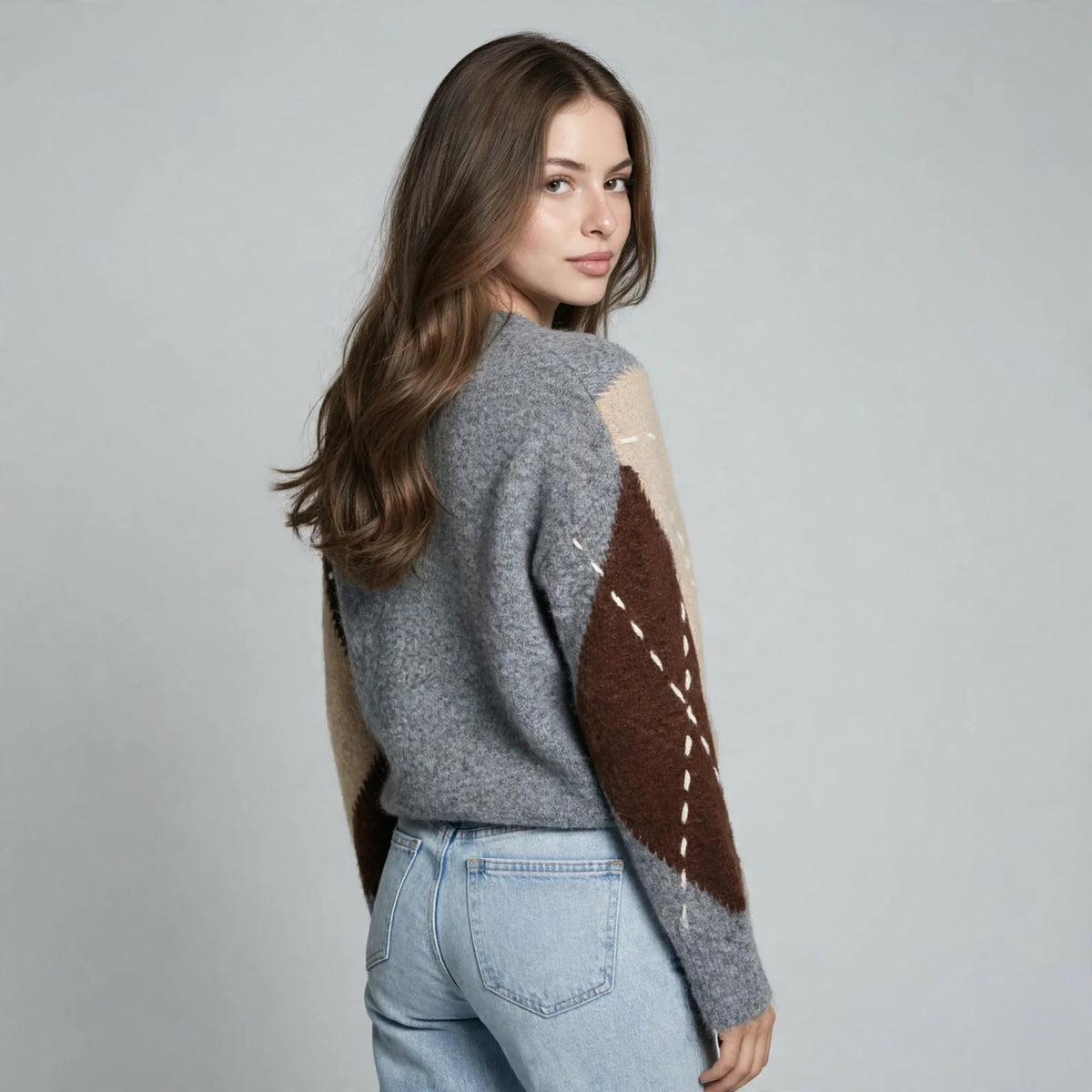 Back three-quarter view of a woman in a gray argyle V-neck oversized sweater and blue jeans, gray studio background.