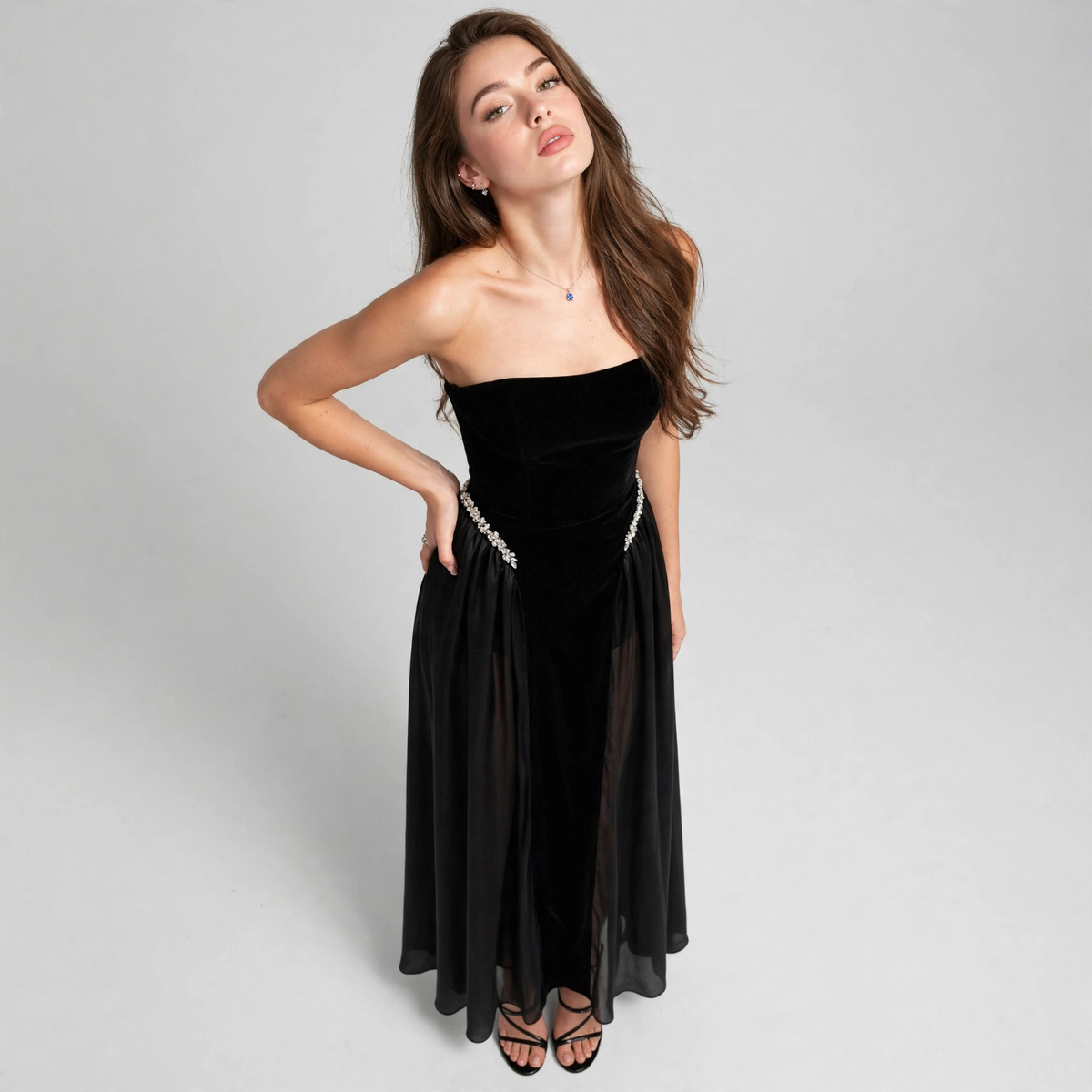 Model leaning to one side wearing black strapless maxi dress with tube bodice and flowing skirt, grey studio background