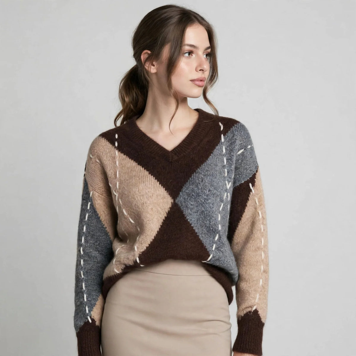 Woman in a brown argyle V-neck oversized sweater, three-quarter pose looking aside, beige pants, gray studio background.