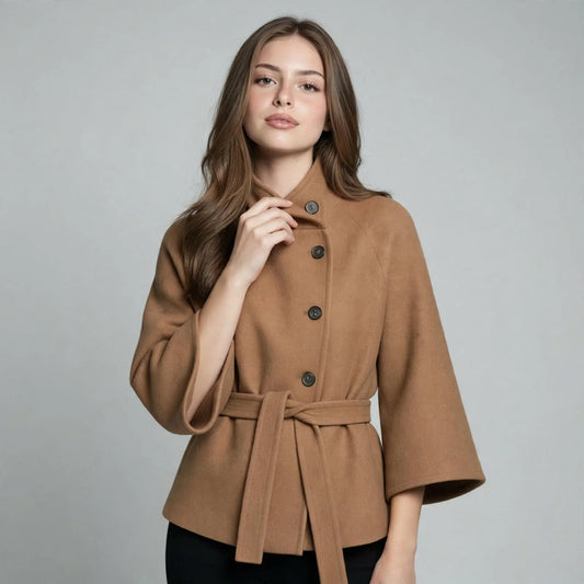 Woman wearing a camel button-down cape coat with belted waist, hand at collar, black pants, gray studio background.