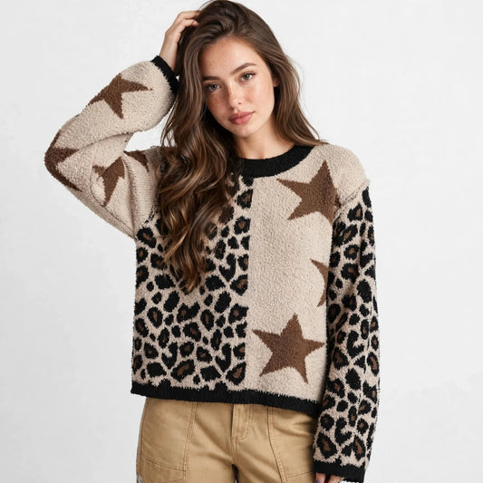Woman with hand in hair wearing a cream knit sweater with brown stars and leopard panels and tan pants, gray studio backdrop.