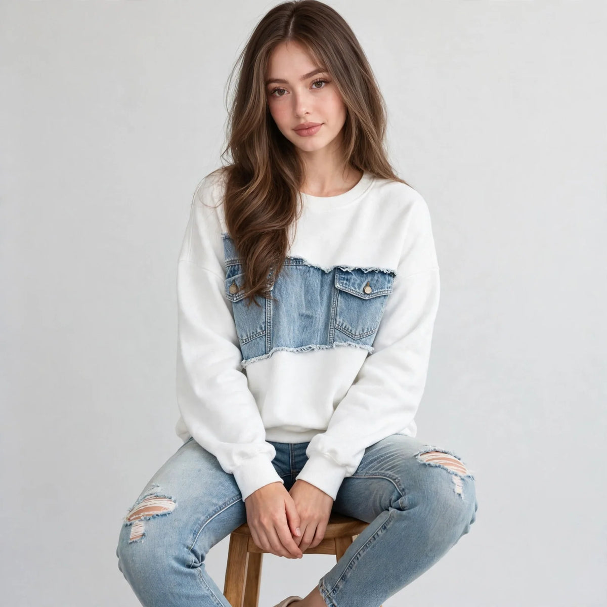 Woman seated on a stool wearing a white denim-patchwork dropped-shoulder sweatshirt and ripped jeans, gray background.