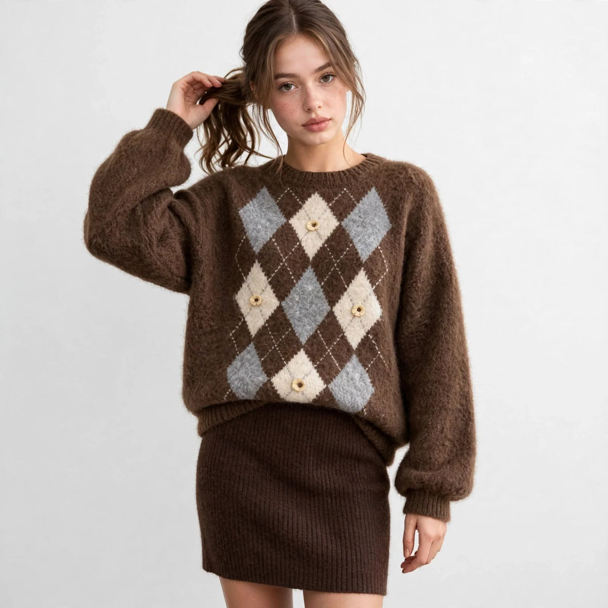 Model with hand in hair in a brown argyle raglan sleeve sweater with cream diamond pattern, brown skirt, white background.