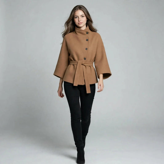 Full-body view of a woman walking in a camel button-down cape coat with belt, black pants, gray studio background.
