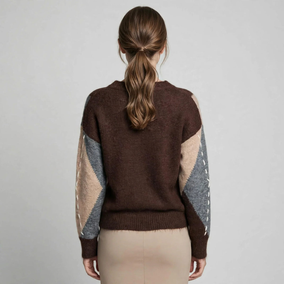 Back view of a woman in a brown argyle V-neck oversized sweater with beige pants, gray studio background.