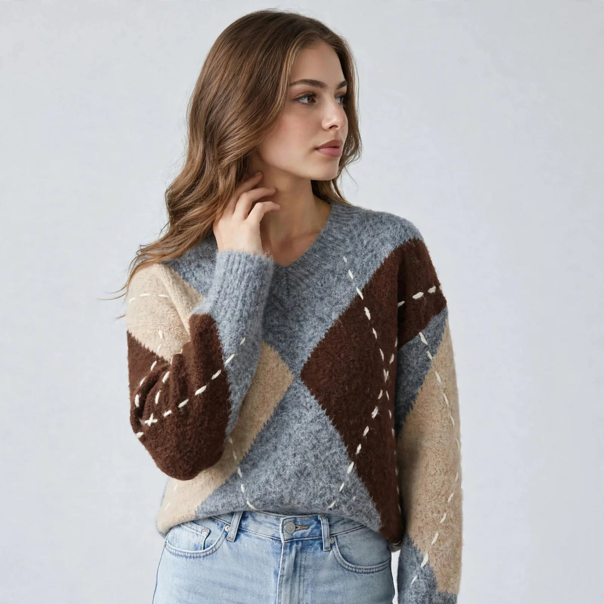 Woman in a gray argyle V-neck oversized sweater with brown and beige diamonds and blue jeans, hand at neck, gray background.