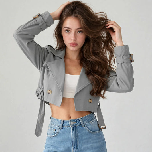 Model in a gray cropped double-breasted jacket over a white crop top and jeans, hands in hair, light gray studio background.