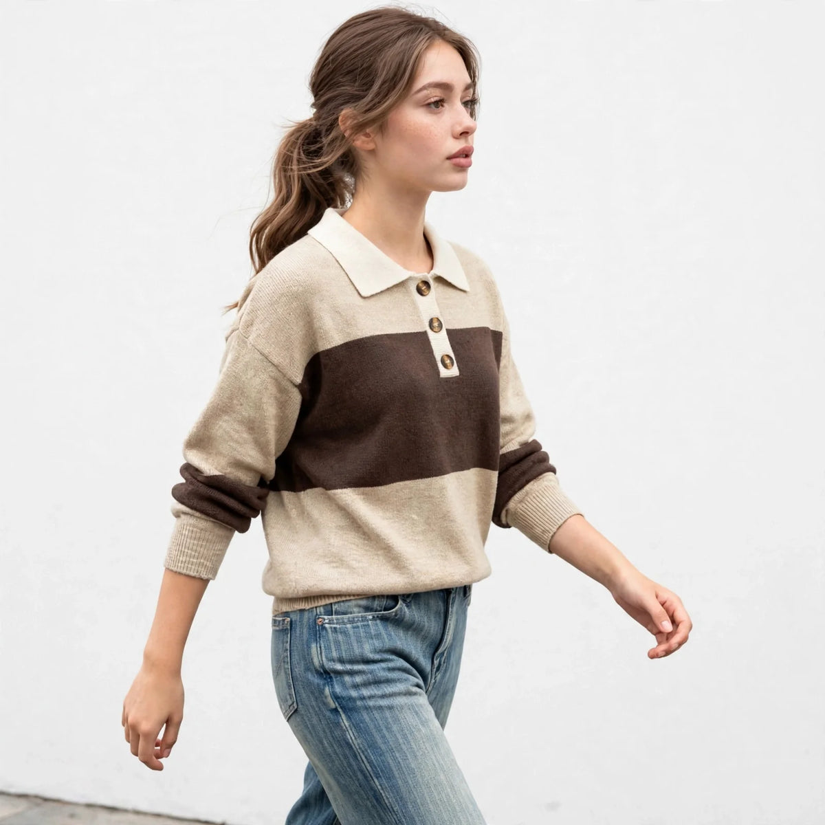 Three-quarter full view of woman walking in beige and brown color-block polo sweater, blue jeans, light studio backdrop.