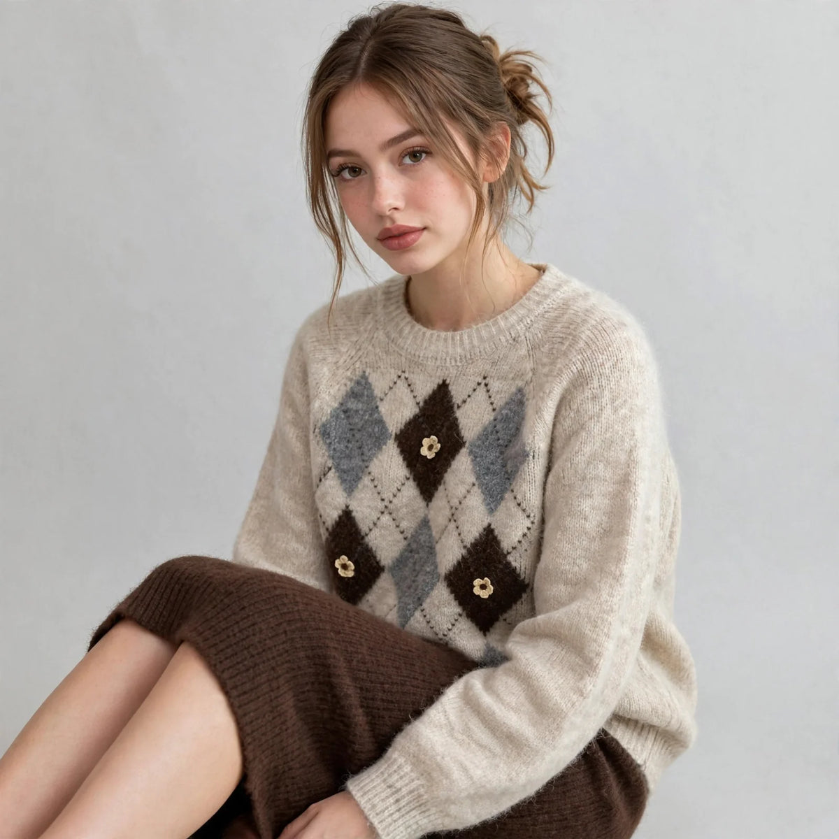 Seated close-up of a beige argyle knit sweater with raglan sleeves and brown diamond pattern, white studio backdrop.
