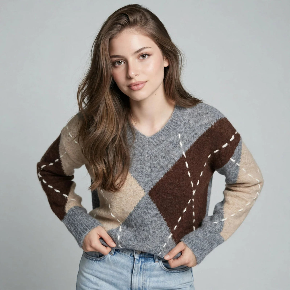 Woman in a gray argyle V-neck oversized sweater with brown and beige diamonds, hands at waist, gray studio background.