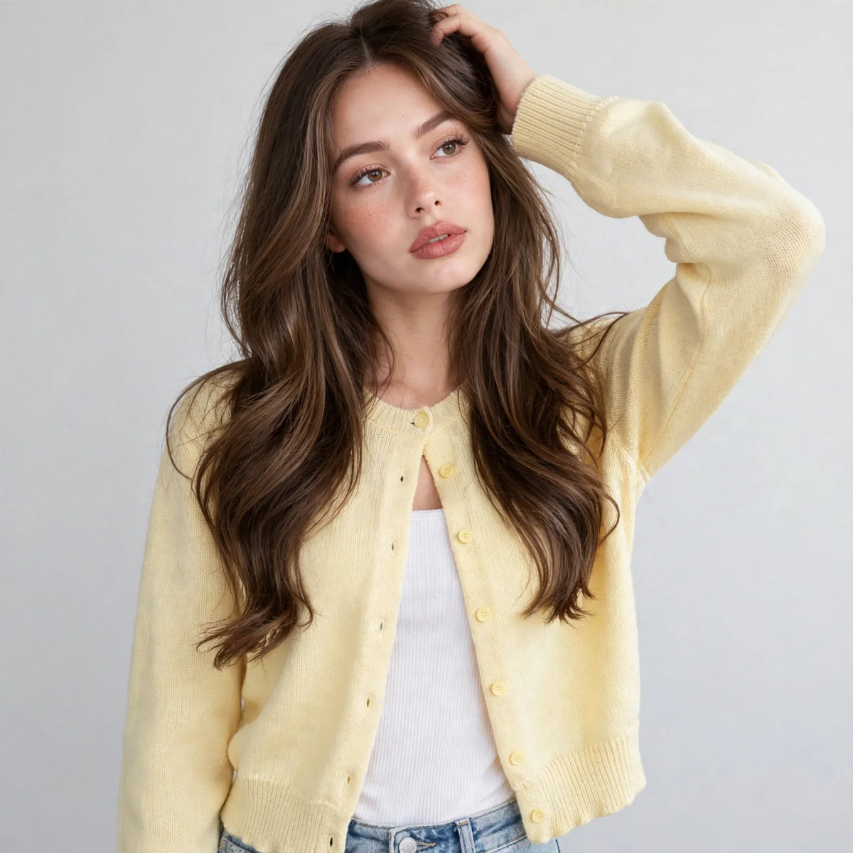 Close-up of model with hand in hair in a pale yellow button-up knit cardigan over a white top, front view, grey background