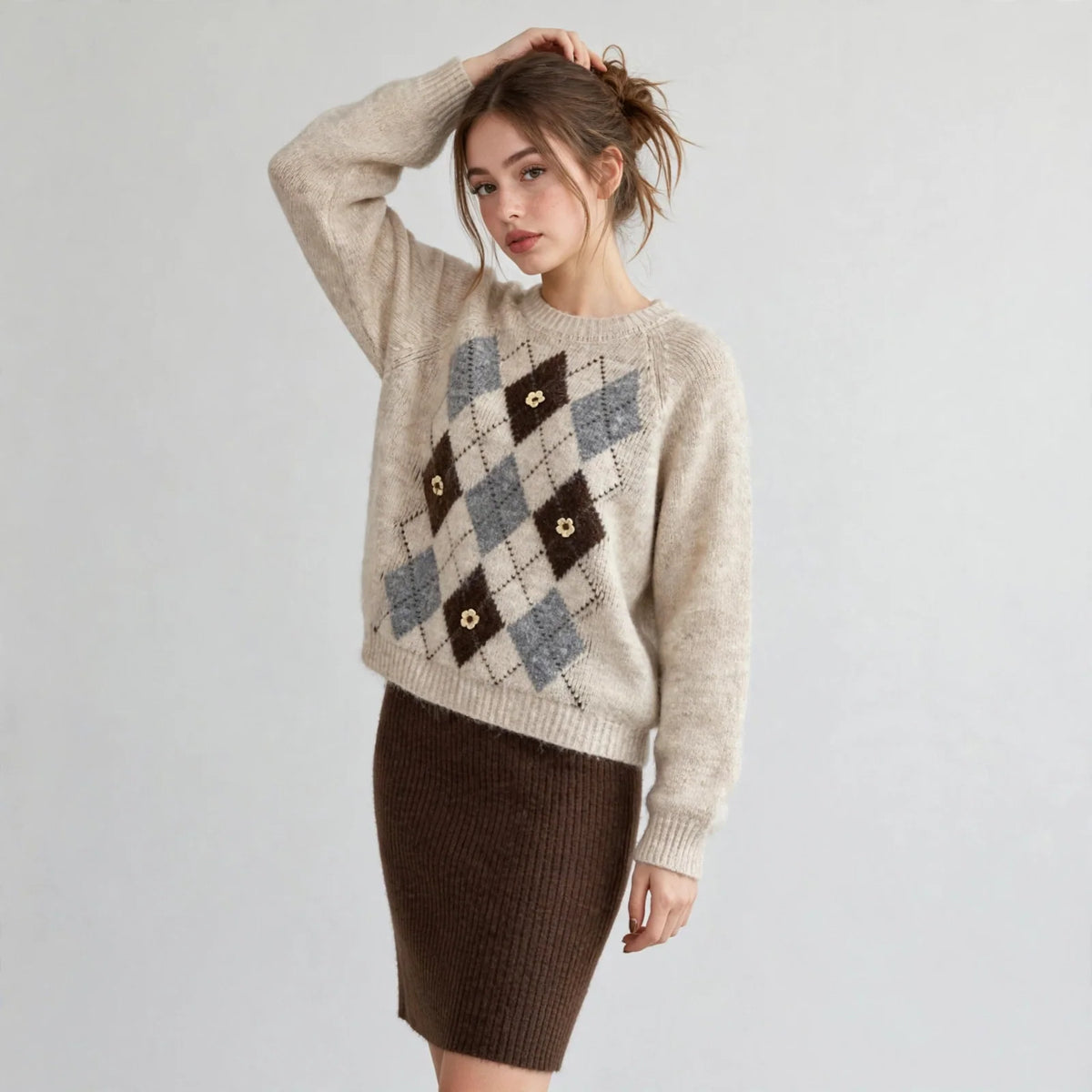 Model with hand in hair wearing a beige argyle raglan sleeve sweater with brown diamond pattern, white studio background.