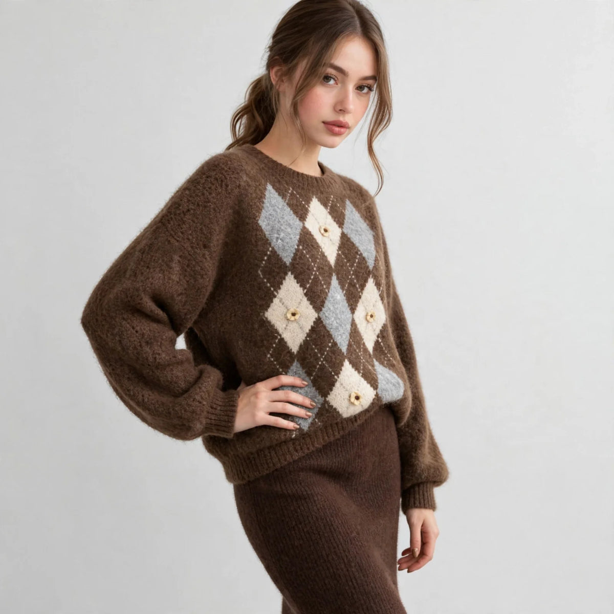 Front three-quarter view of a brown argyle knit sweater with raglan sleeves, hand on hip, brown skirt, white studio backdrop.