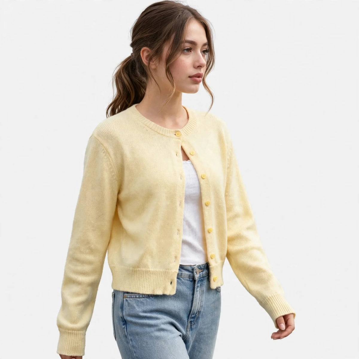 Front three-quarter view of model walking in a pale yellow button-up knit cardigan over a white top and blue jeans