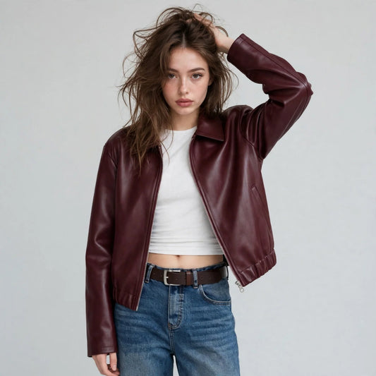Woman, hand in hair, in burgundy faux leather zip-up jacket, shirt collar, white top, jeans, front view, grey backdrop.