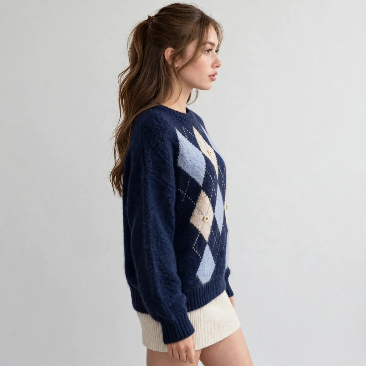 Side profile of a navy argyle raglan sleeve sweater with light blue and cream diamonds, white skirt, white studio backdrop.