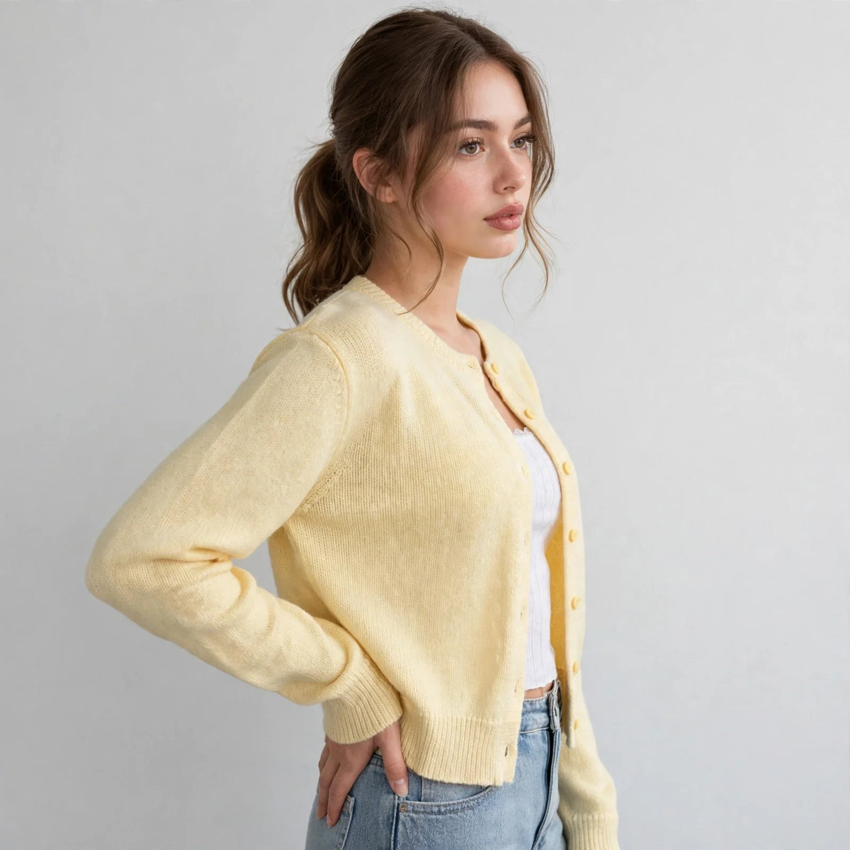 Side view of model with hand on hip wearing a pale yellow button-up knit cardigan with long sleeves and blue jeans