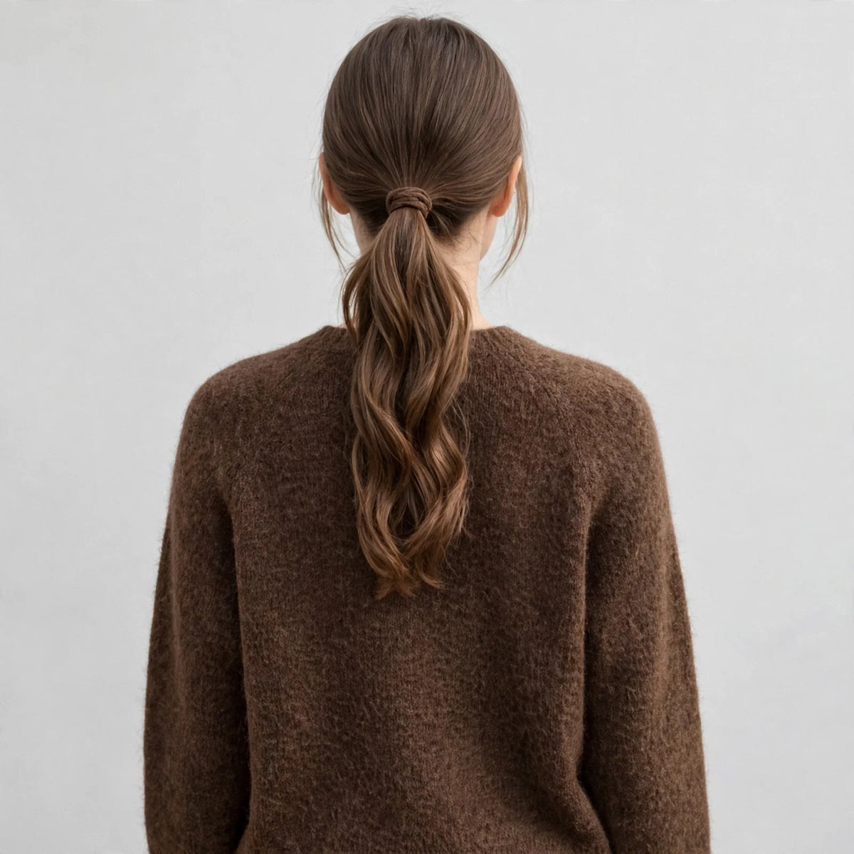 Back view of a brown argyle raglan sleeve knit sweater, model with low ponytail, white studio background.