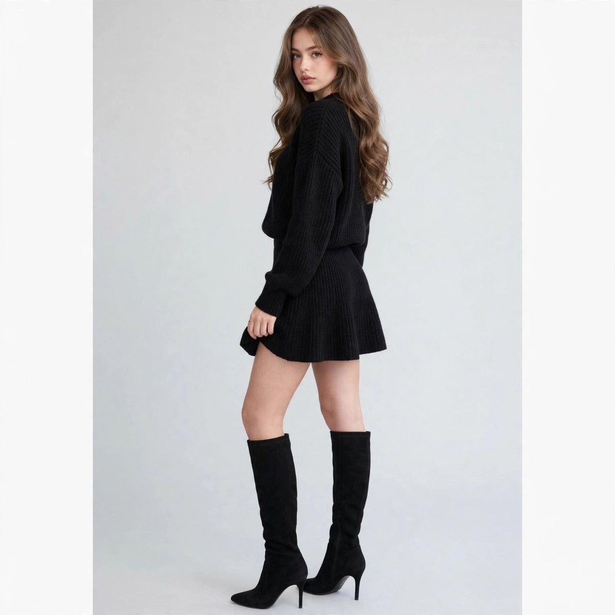 Back three-quarter view of a black ribbed knit sweater and flared mini skirt set with black knee-high boots, gray studio.