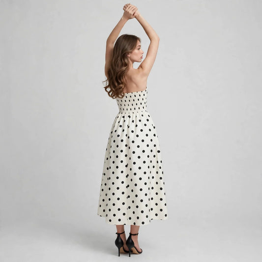 Back view of white polka dot strapless midi dress with black dots, smocked bodice, model with arms raised, studio backdrop.