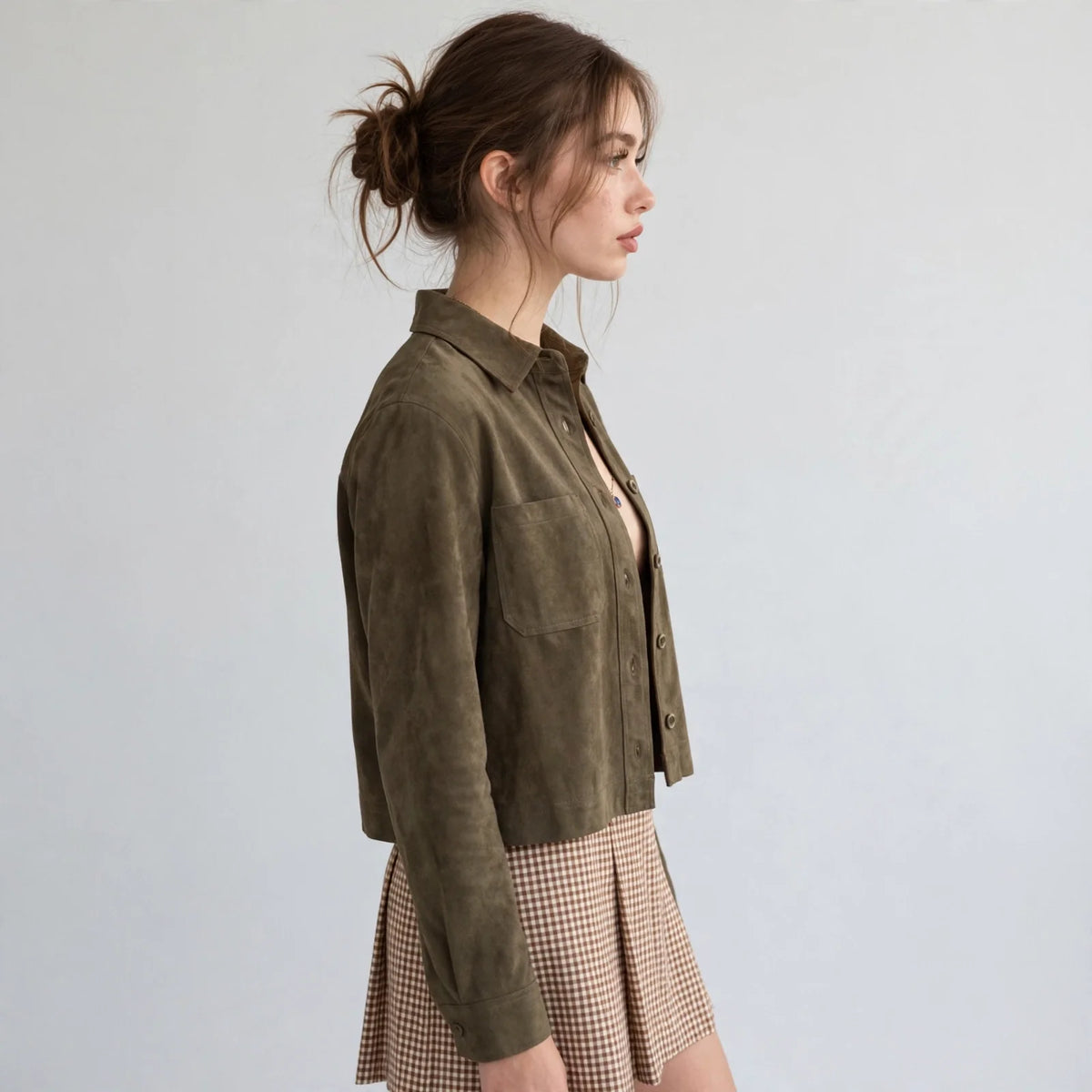 Woman in side profile wearing an olive cropped faux suede button-down jacket and a checkered skirt, gray background.