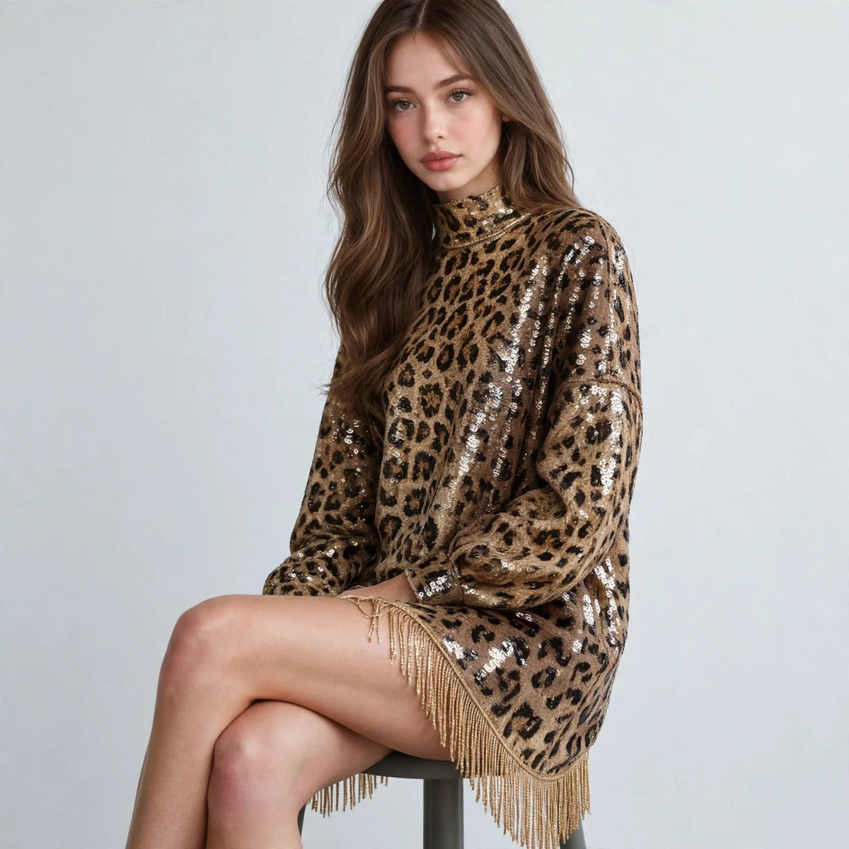 Seated model wearing a leopard sequin mock neck mini dress with long sleeves and fringe hem, black stool, white background.