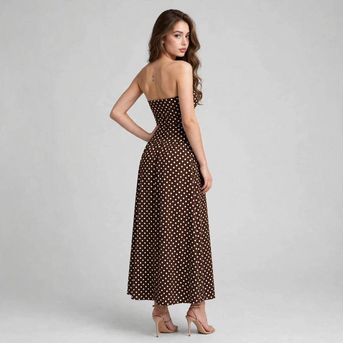 Back three-quarter view of a brown polka dot strapless maxi dress with smocked bodice, hand on hip, white backdrop.